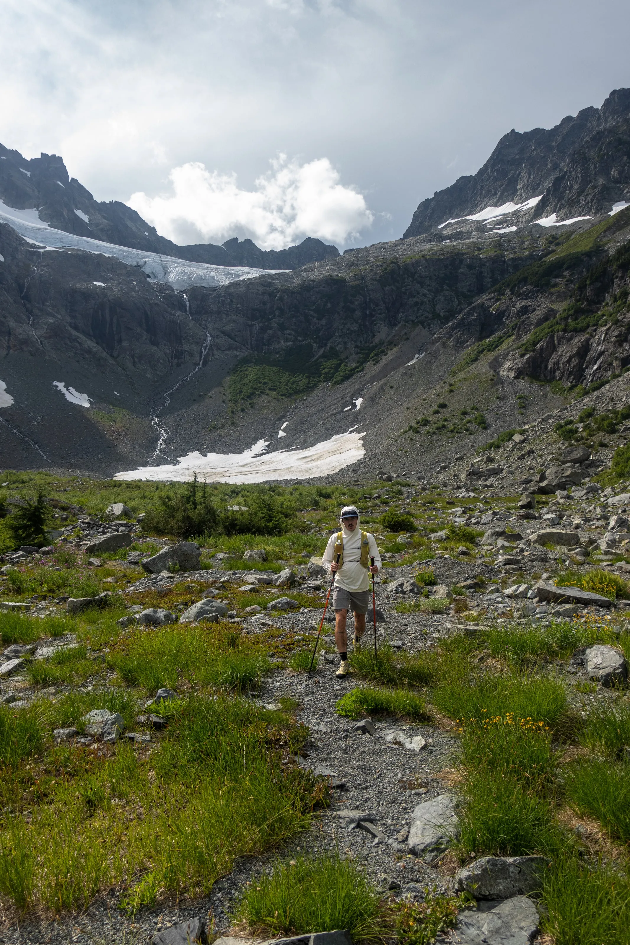 A climber walking with trekking poles through rocky subalpine terrain at Russian Army Camp below a glacier and dark ridgelines