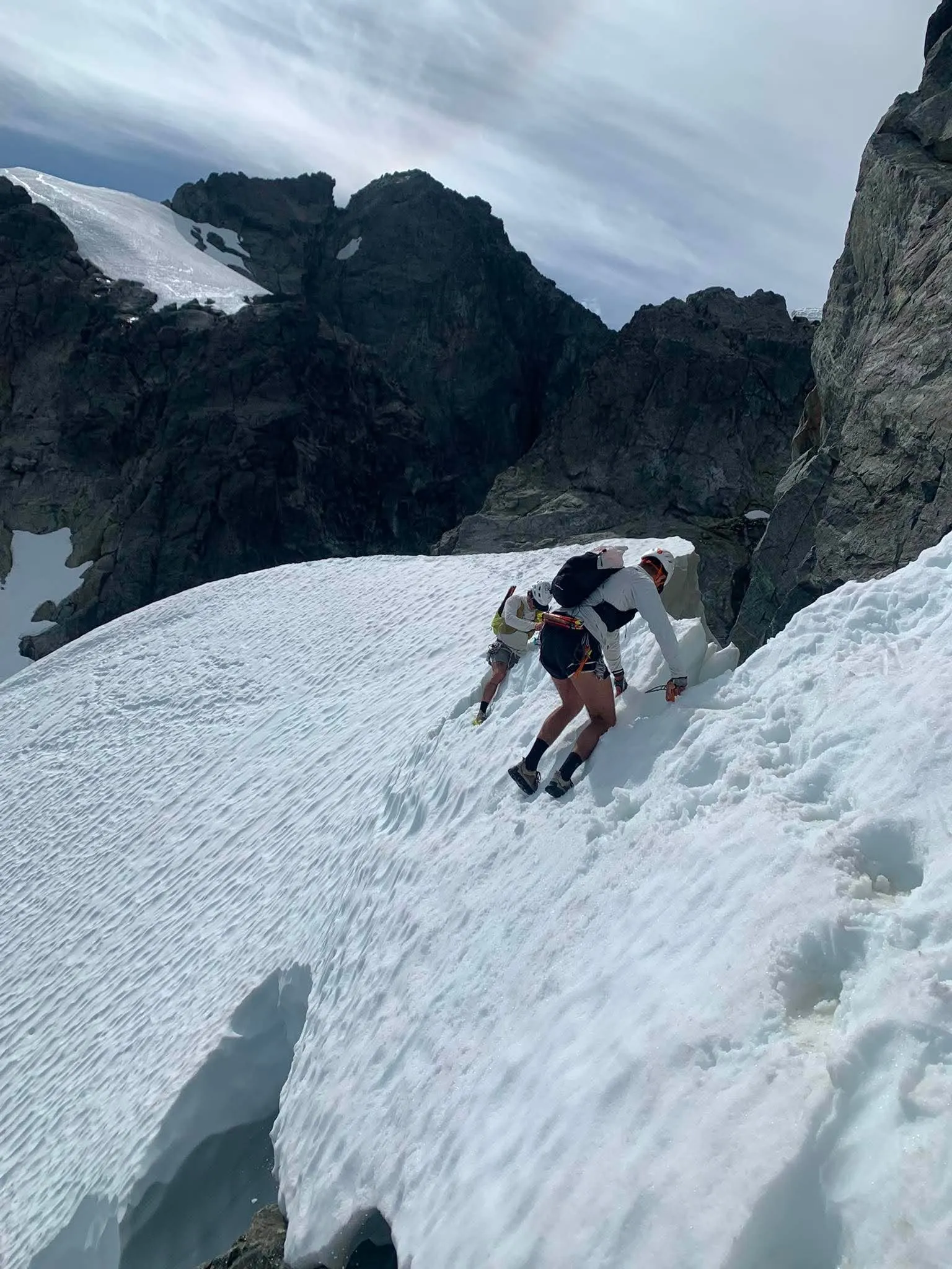 Two climbers wearing helmets and crampons moving carefully across steep summer snow between rock walls